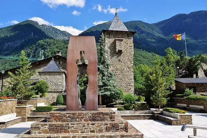 Historic stone architecture in Andorra with a modern sculpture, surrounded by lush greenery and mountain views.