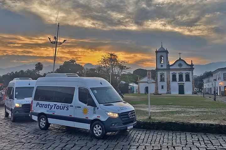 Sunset view of a Paraty Tours van parked on cobblestone street in Angra dos Reis, Brazil, near a historic church.