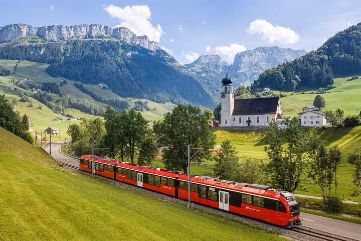 Scenic railway journey through Appenzell with a red train passing by a picturesque church and lush green mountains.