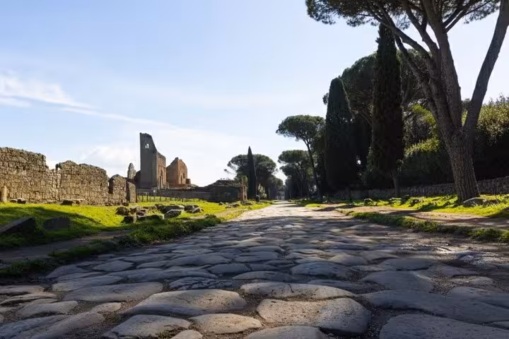 Ancient cobblestone road along the Appian Way, lined with ruins and cypress trees under a clear blue sky.