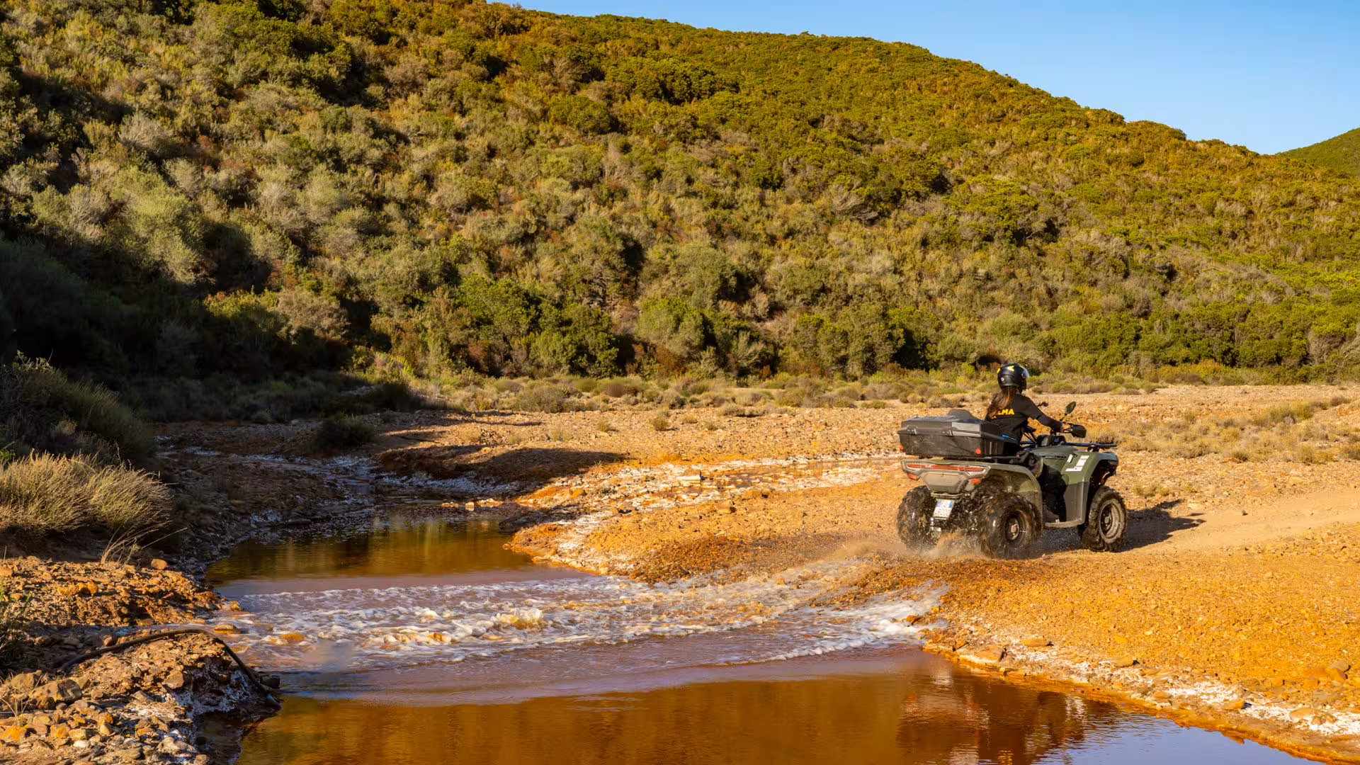 Adventurer on ATV crossing scenic stream in Costa Verde during exciting Arbus off-road excursion.