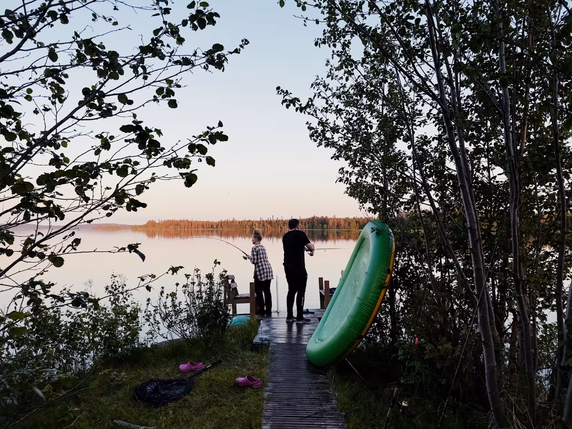 Two people fishing at sunset on a serene Arctic lake in Rovaniemi, surrounded by lush greenery and calm waters.