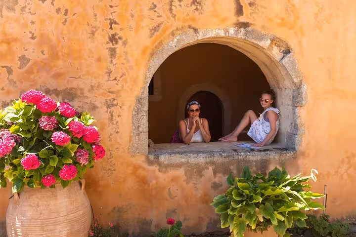 Two people relax in a rustic stone window framed by vibrant flowers on a sunny day in Arles, perfect for a city tour.