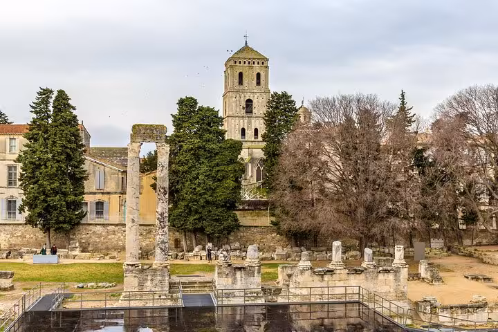 Ancient Roman ruins with a historic church tower and lush trees in Arles, perfect for a cultural city walk.