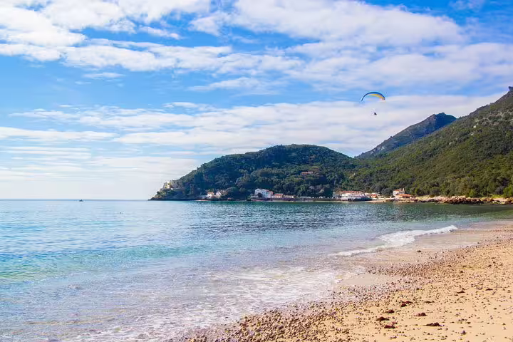 Scenic Arrábida coastline with sandy beach, clear blue waters, lush hills, and a paraglider soaring under a sunny sky.