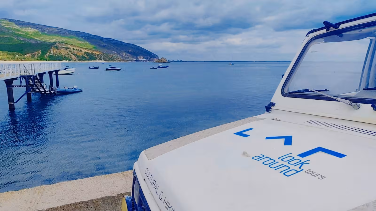 4x4 jeep by Setúbal bay pier on Arrábida Safari Tour, with calm sea, boats and green mountains in background