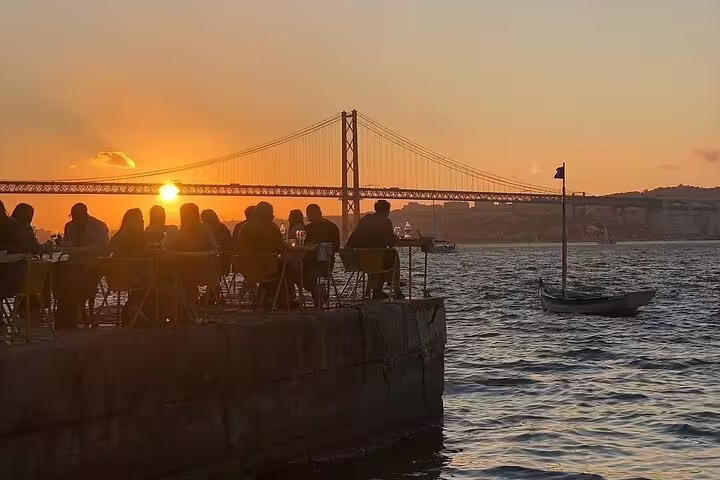 People enjoy a scenic sunset by the waterfront with a view of the bridge on the Arrábida & Sesimbra day trip, featuring wine tasting and nature exploration.