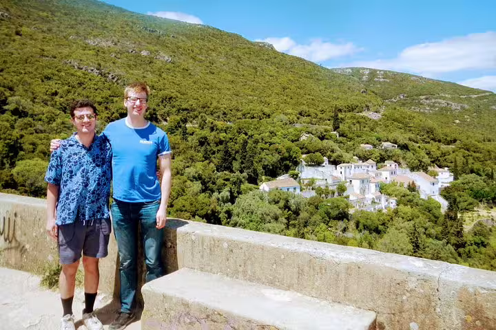 Two people stand on a scenic viewpoint in Arrábida, overlooking lush green hills and a picturesque village under a clear blue sky.