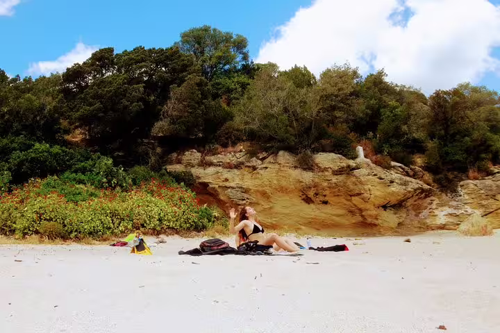 A woman relaxes on a sandy beach with rocky cliffs and lush greenery in Arrábida, ideal for a snorkeling and wine tour experience.