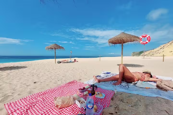 Relaxing beach picnic setup with red checkered blanket, sunbathing under straw umbrellas on Arrábida's scenic sandy shores.
