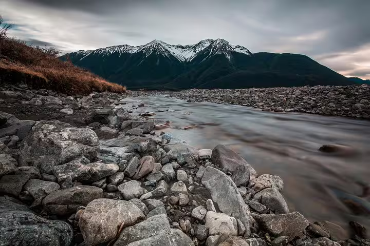 Serene river flowing through rocky terrain with snow-capped mountains in Arthur's Pass, ideal for a scenic day tour.