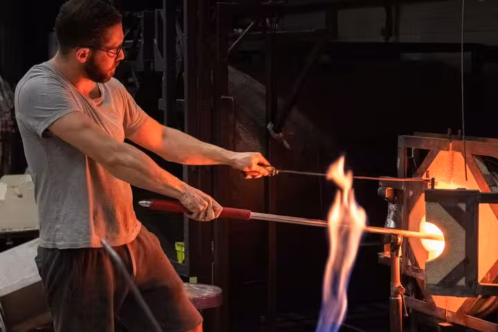 Artisan glassblower skillfully shaping molten glass amidst fiery furnace, showcasing traditional craftsmanship techniques.