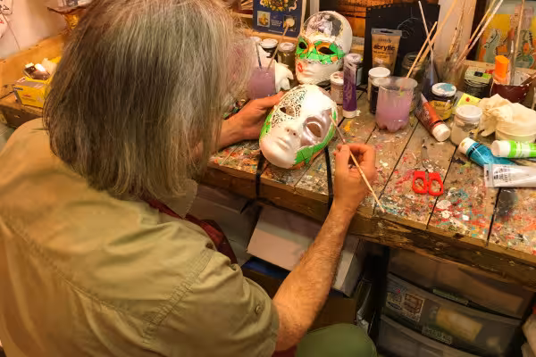 Close-up of artisan applying delicate details to a mask, with an array of colorful paints and crafted masks nearby.