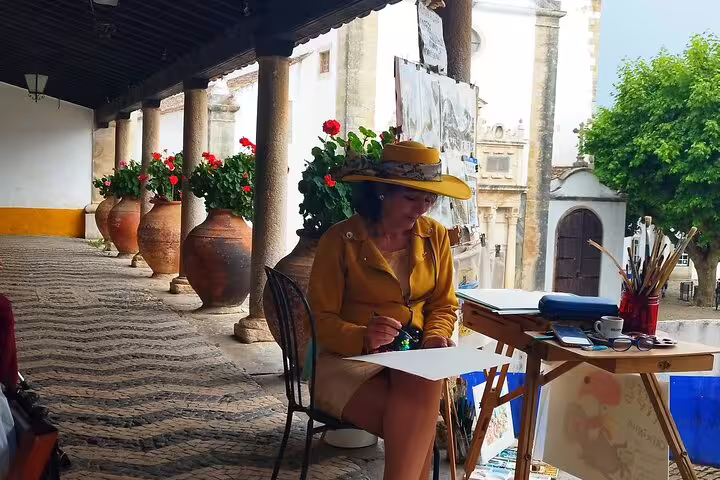 Artist painting on a scenic terrace in Obidos, surrounded by vibrant flowers and historic architecture, during a Lisbon day tour.