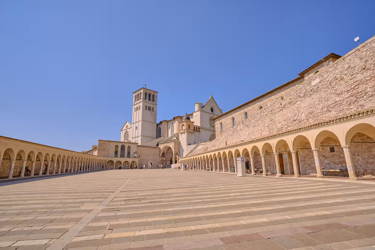 Wide-angle view of Assisi's Basilica with its grand architecture and serene courtyard under a clear blue sky.