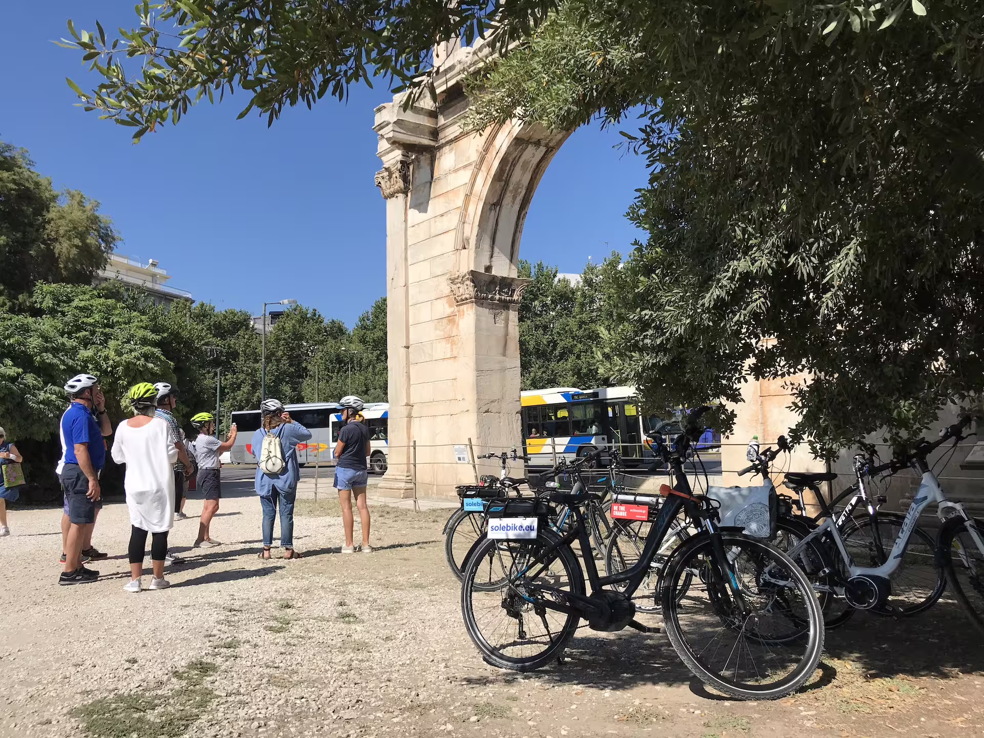 Tourists on e-bikes explore Athens' ancient landmarks near the Arch of Hadrian under a sunny blue sky.