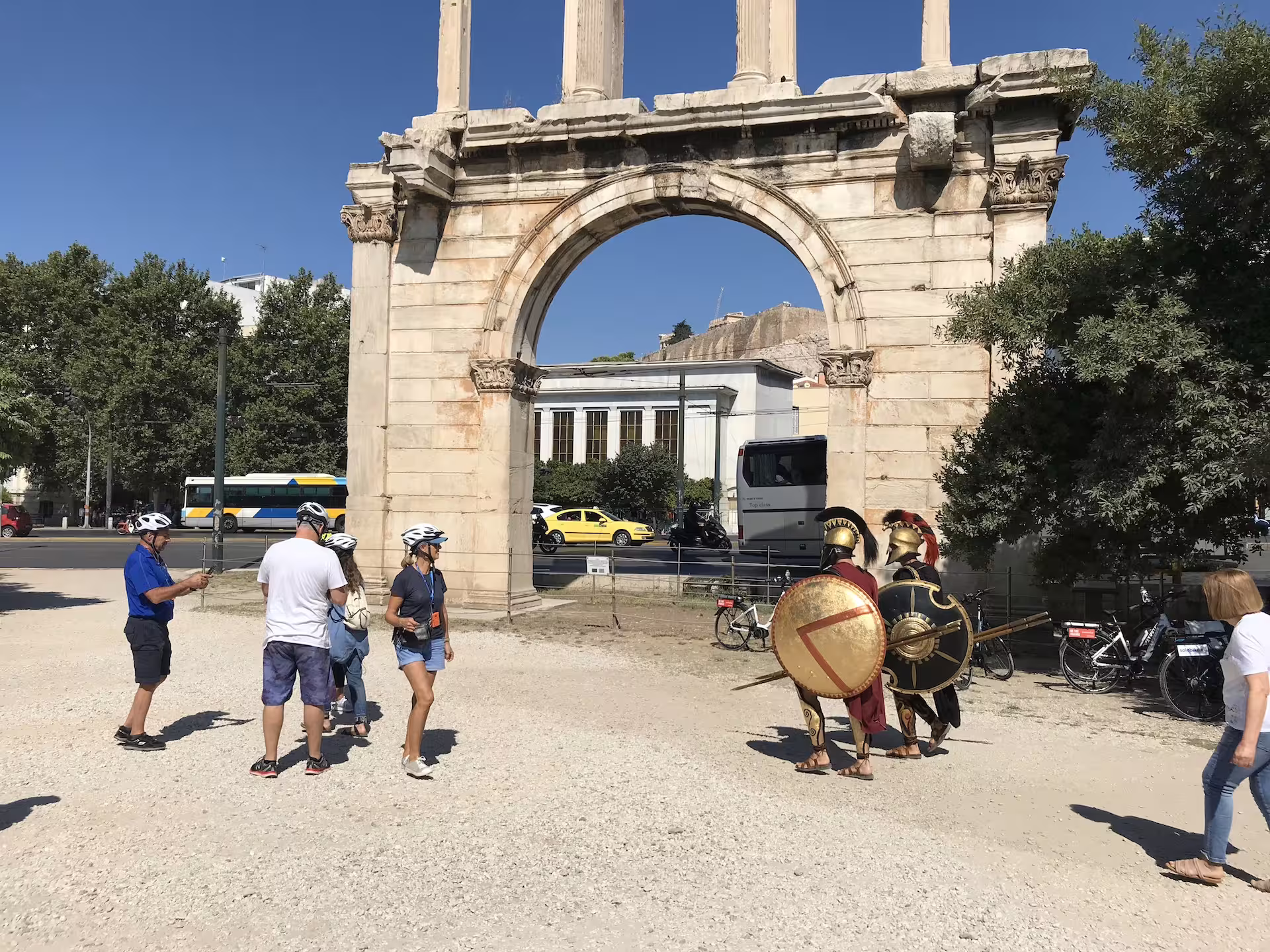 Visitors on e-bikes admire the Arch of Hadrian in Athens, with actors dressed as ancient Greek warriors nearby.