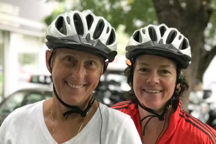 Two smiling cyclists wearing helmets, ready for an Athens e-bike tour with a backdrop of green trees.