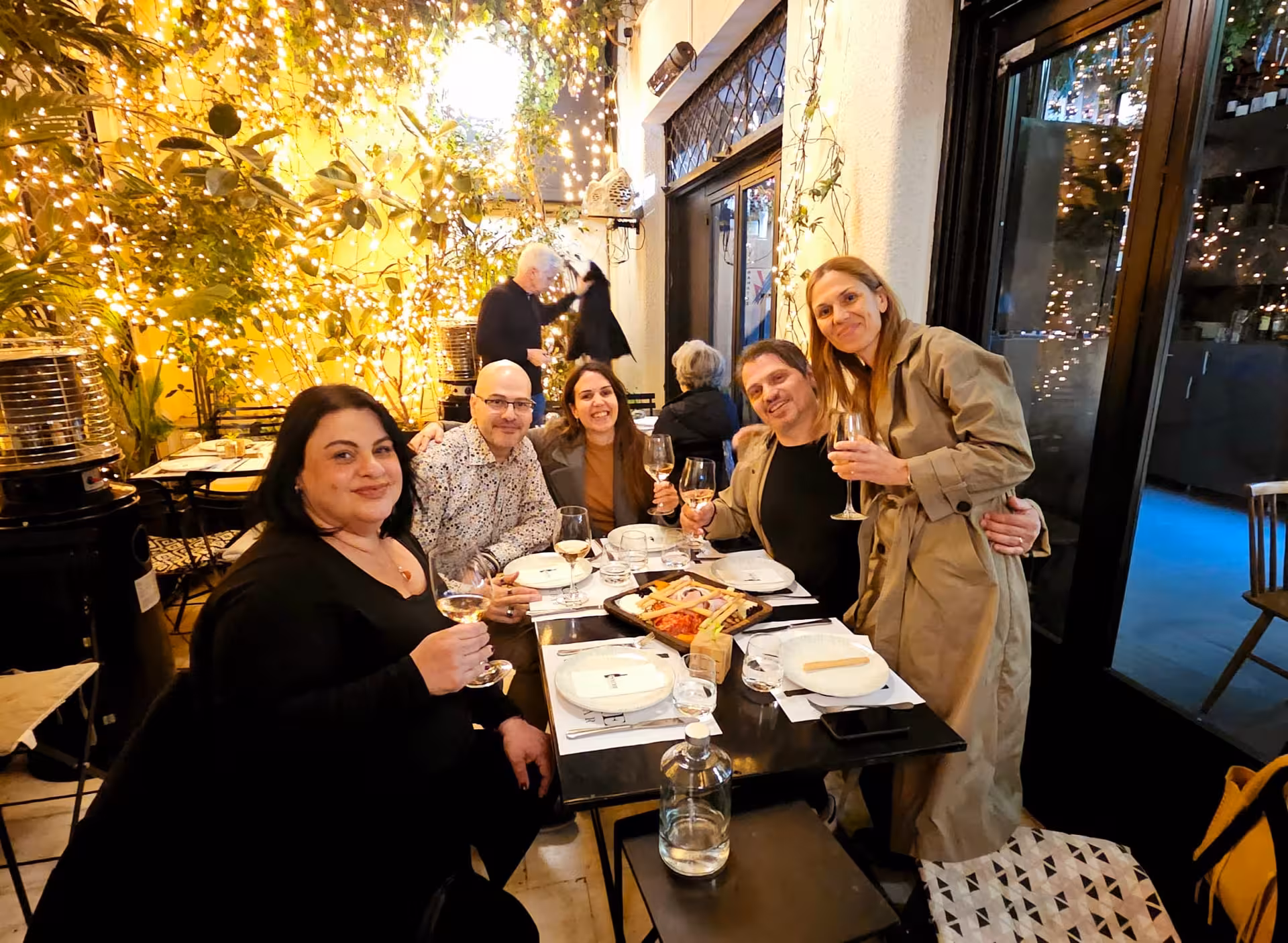 Friends toast Greek wine at a cozy Athens evening wine tour, enjoying meze under twinkling courtyard lights