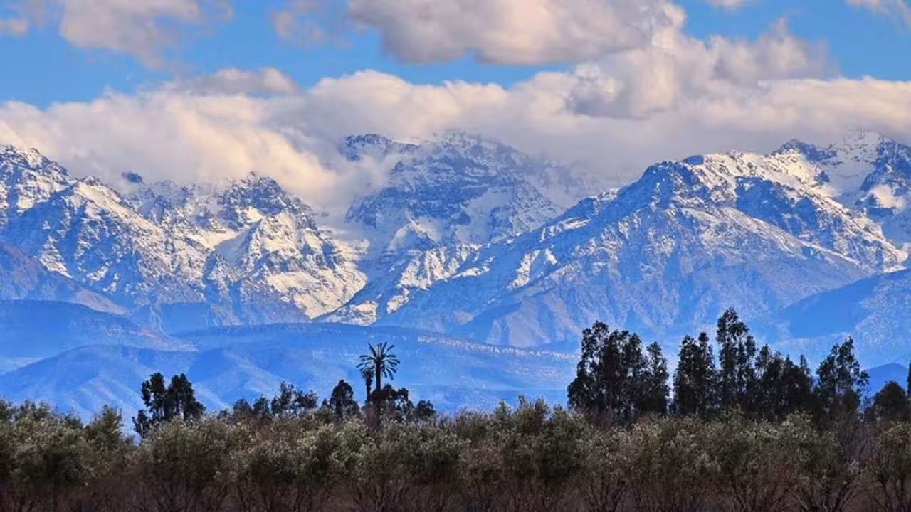 Majestic snow-capped Atlas Mountains with lush greenery and clouds in the foreground near Marrakech.