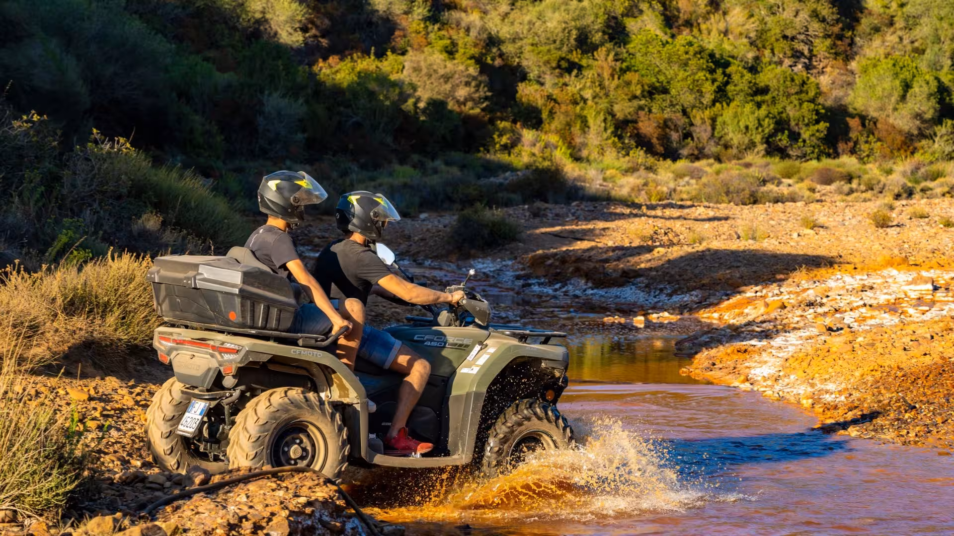 Two riders navigating an ATV through a rocky stream, surrounded by the vibrant greenery of Costa Verde.