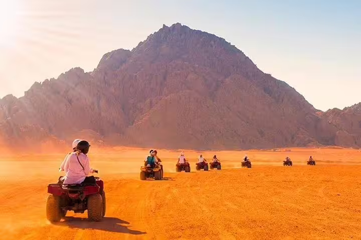 Group riding ATV quad bikes across Hurghada desert toward mountains at sunset, with dinner and Bedouin show