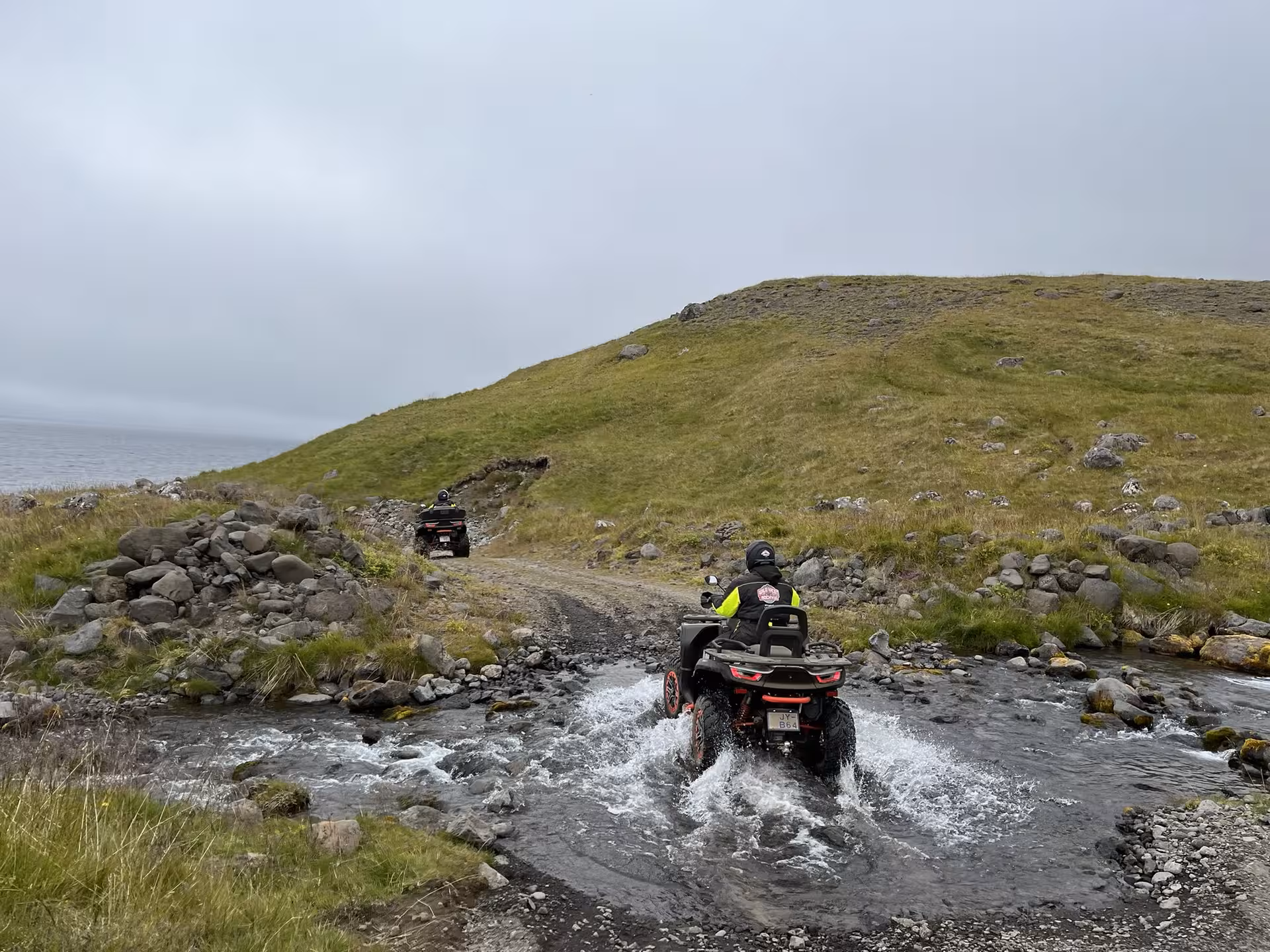 ATV tour navigates through a scenic water crossing on the rocky Svalvogar trail under an overcast sky.