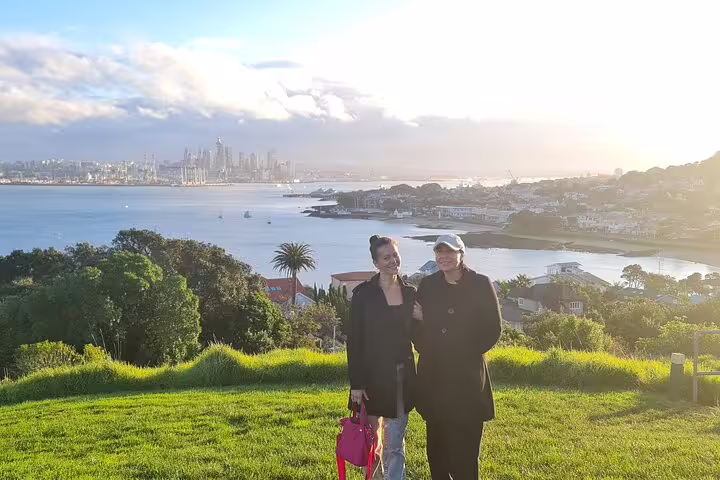 Two people enjoying a scenic view of Auckland's skyline and harbor from a lush green hill during sunset.