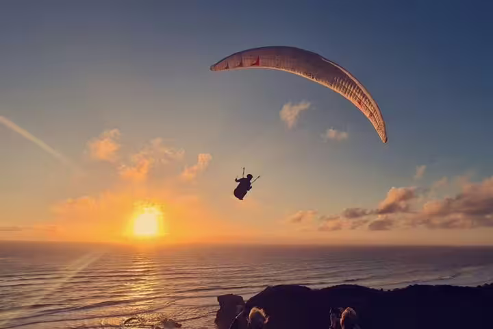 Paraglider soaring against a vibrant Auckland sunset by the ocean, highlighting adventure and romance on a scenic tour.