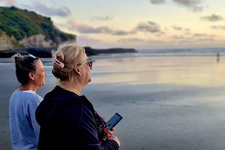 Two women enjoying a tranquil Auckland beach sunset on romantic tour with winery and thermal pool experience.