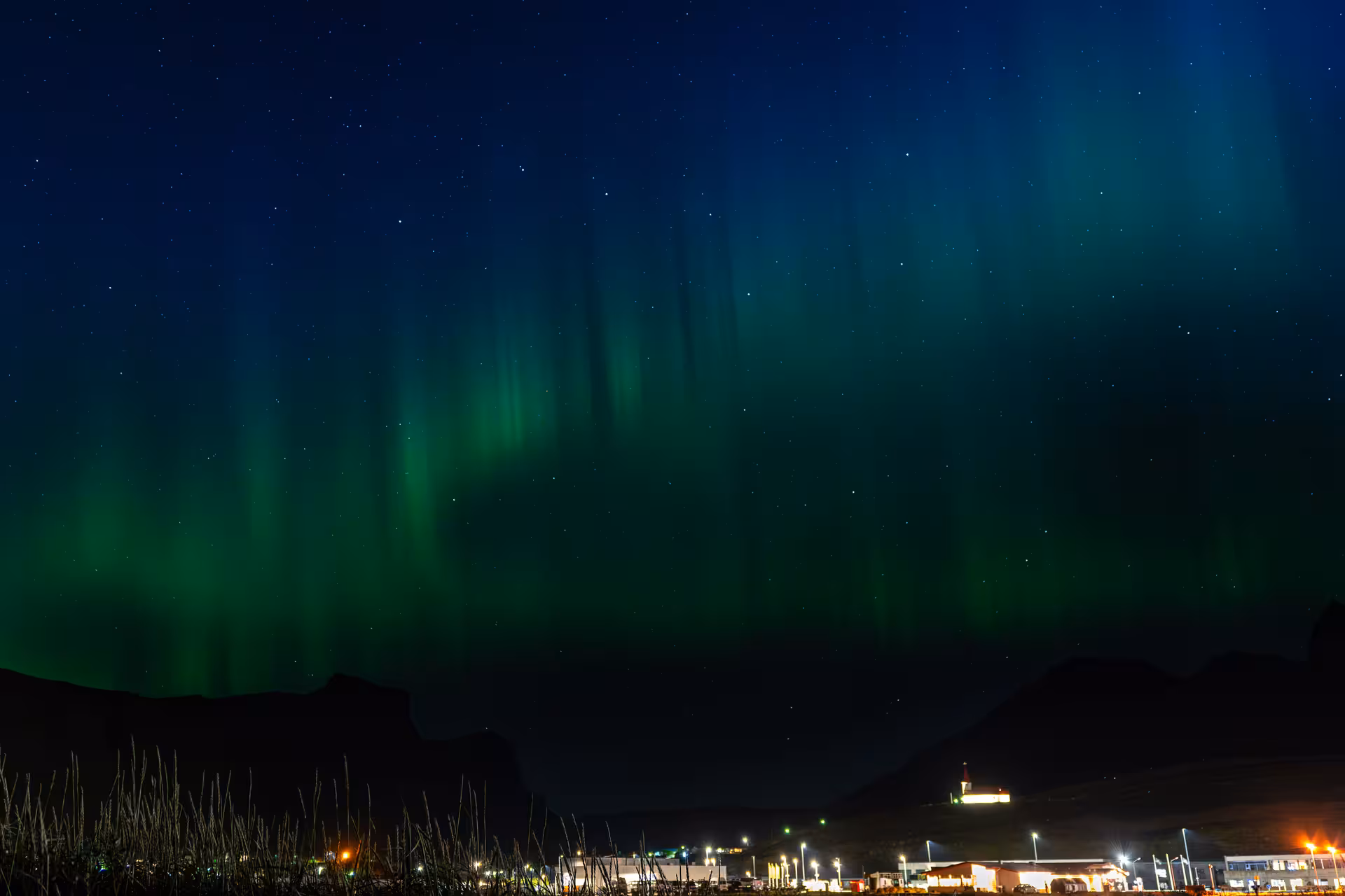 Aurora borealis curtains over Iceland town lights and mountains, Northern Lights tour viewing and photos