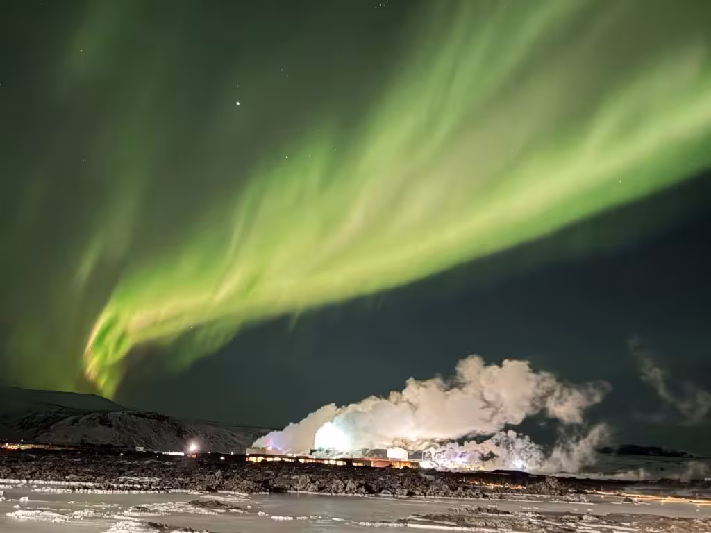 Vivid green aurora borealis over snowy landscape and steaming geothermal plant, Iceland northern lights tour