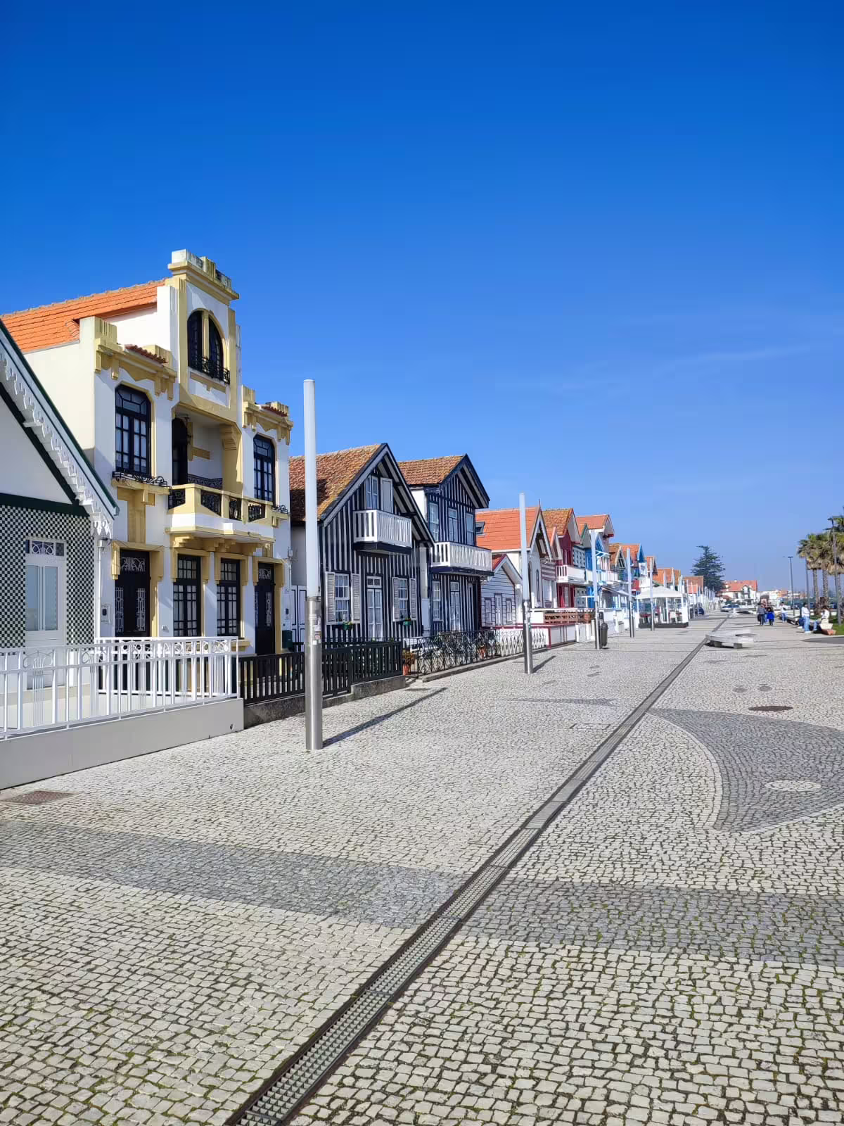Colorful traditional houses line a cobblestone street in Costa Nova, Aveiro, under a clear blue sky.