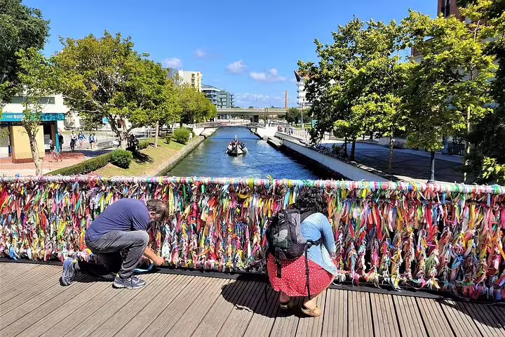 Tourists attach colorful ribbons to a bridge in picturesque Aveiro, Portugal, with a canal and boat in the background.