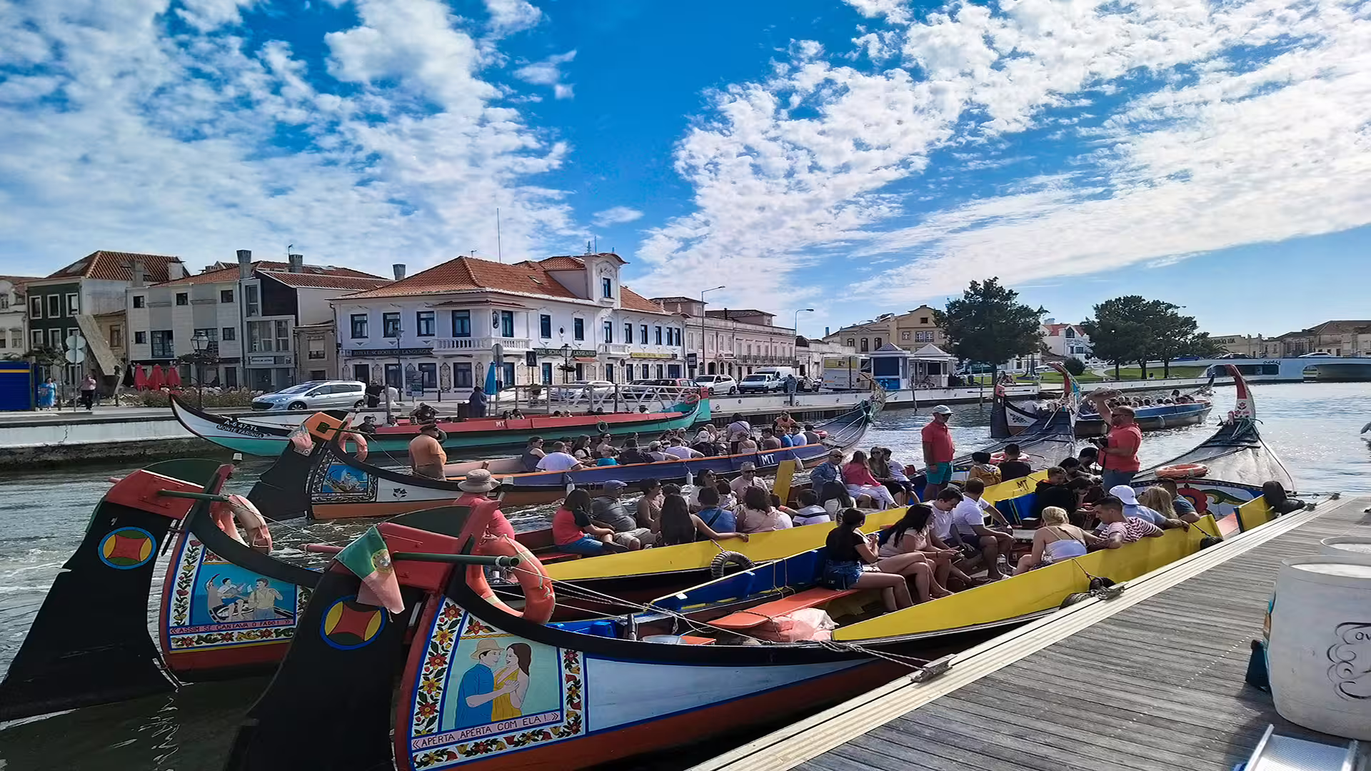 Tourists enjoy a scenic boat ride in Aveiro, Portugal, with colorful moliceiro boats and charming architecture under a vibrant sky.