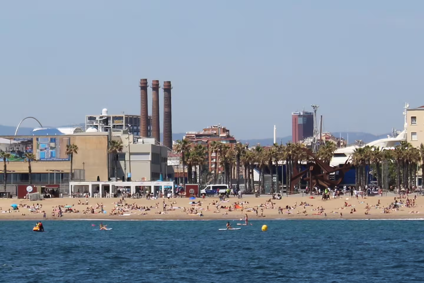 Sunny beach in Barcelona bustling with visitors, framed by palm trees and urban architecture.