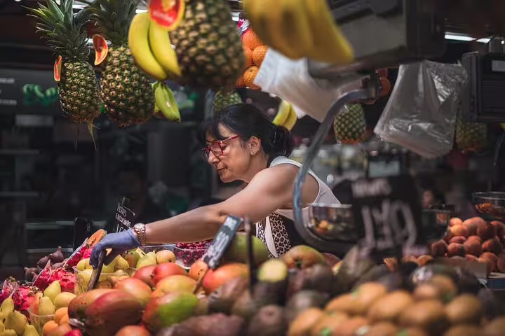 A woman selecting fresh produce at a vibrant market, perfect for sourcing ingredients for the Ultimate Paella Cooking Class Experience.