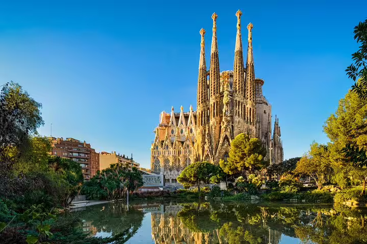 Stunning view of Barcelona's Sagrada Familia against a clear blue sky, reflecting in a serene pond surrounded by greenery.
