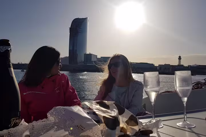 Two women enjoy drinks during a private sunset sailing tour in Barcelona, with a view of the city skyline and waterfront.