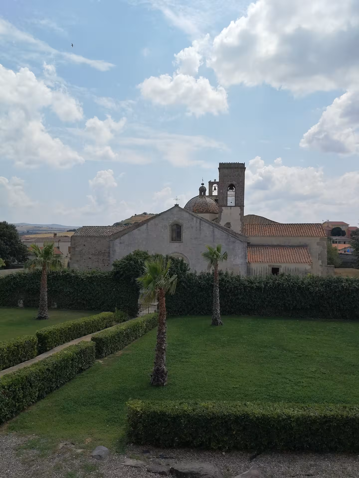 Scenic view of a historic church near Barumini, surrounded by lush gardens under a partly cloudy Sardinian sky.