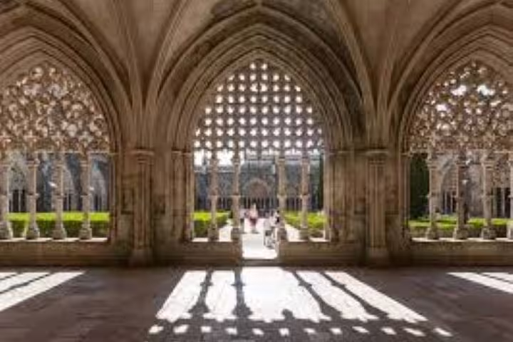 Sunlit Gothic arches casting shadows in Batalha Monastery, with a view of the lush courtyard beyond the intricate stonework.