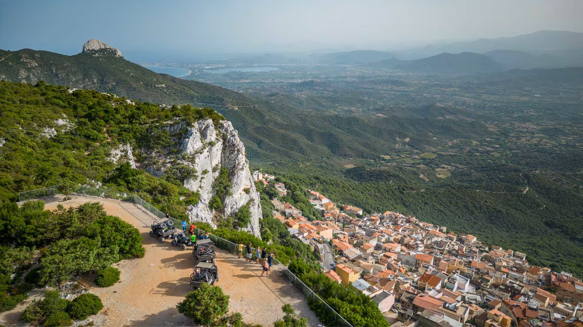 Scenic view from Baunei featuring buggies parked on a cliff overlooking the lush Sardinian landscape and village.