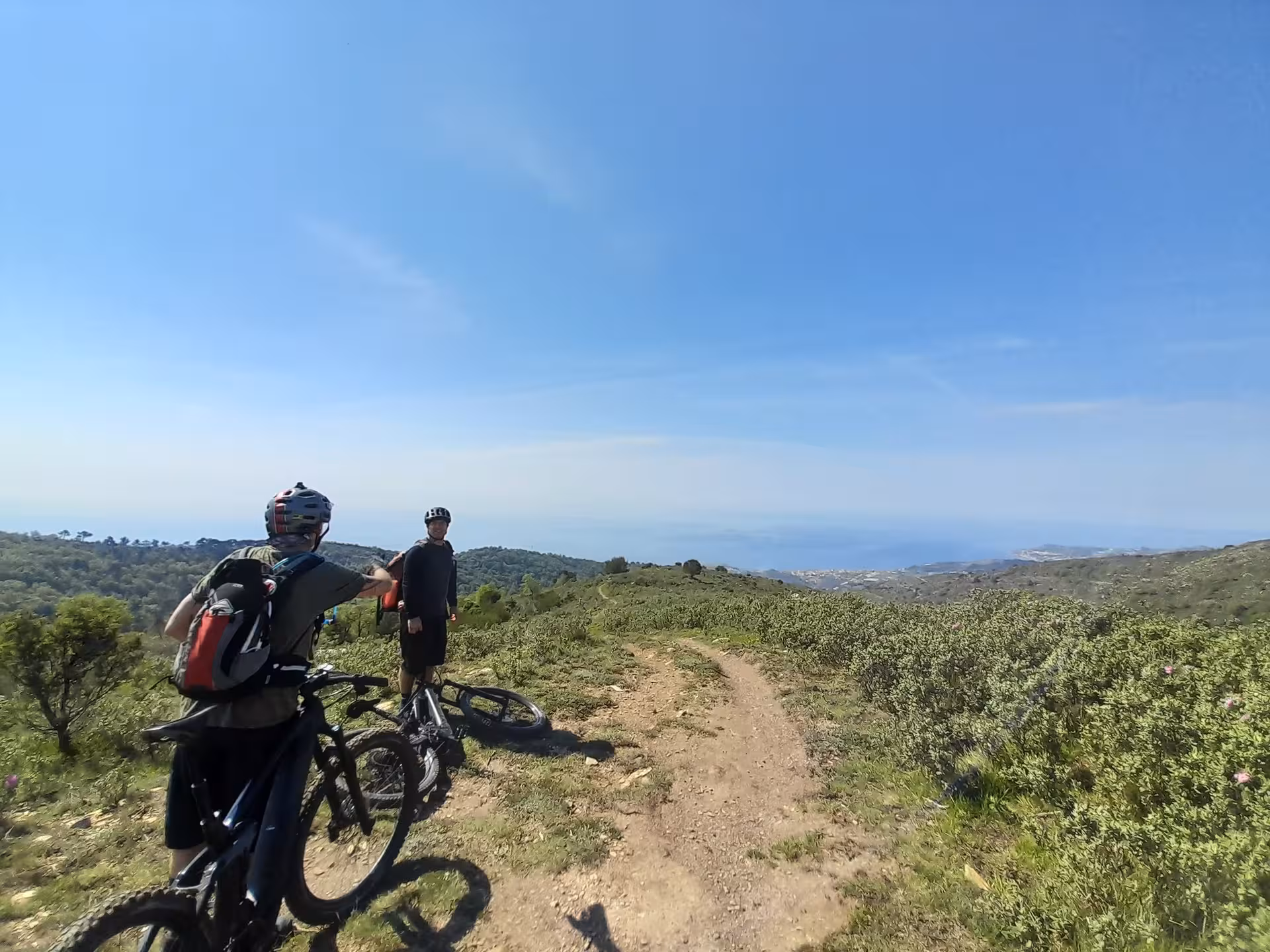 Two mountain bikers pause on a scenic trail overlooking a vast landscape and ocean view, part of a beginner MTB tour.