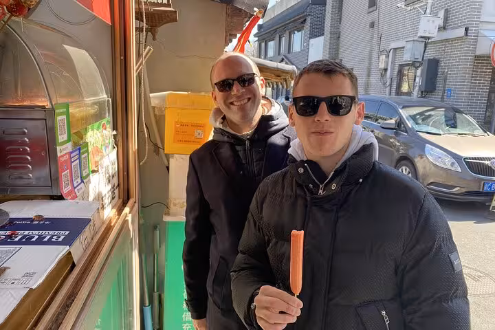 Two friends relish local snacks outside a Beijing street vendor during a vibrant sidecar food tour.