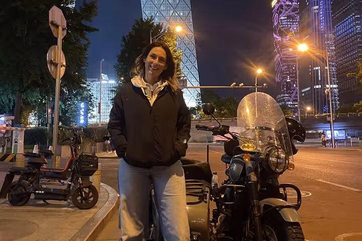 Woman stands beside a sidecar motorcycle, illuminated by vibrant city lights during a night tour in Beijing.