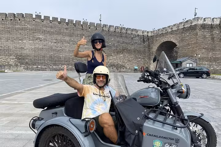 Tourists enjoy a thrilling Beijing sidecar ride, posing with thumbs up near historic city walls on a sunny day.