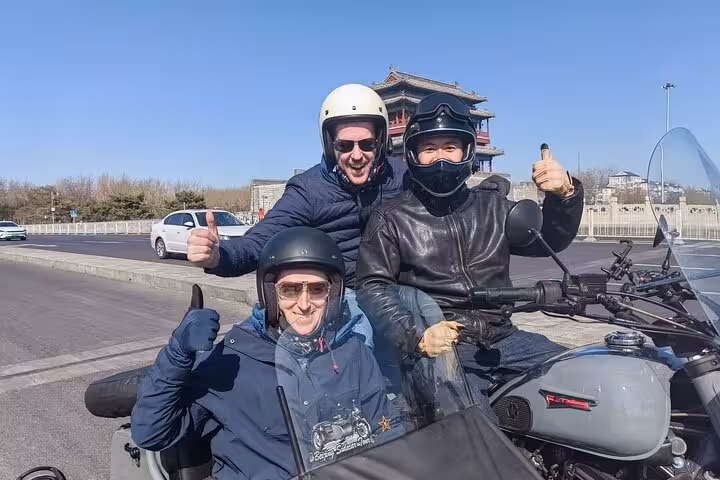 Smiling riders on a Beijing sidecar tour with a historic tower in the background, enjoying a sunny day.
