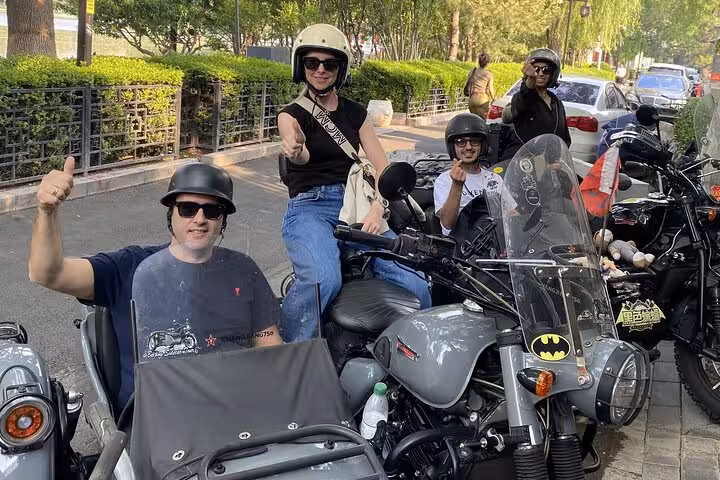 Tourists enjoy a Beijing sidecar tour, giving thumbs up while parked beside vibrant city streets.