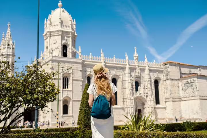 A traveler with a backpack and hat admires the historic architecture of Jerónimos Monastery in Belém on a sunny day.