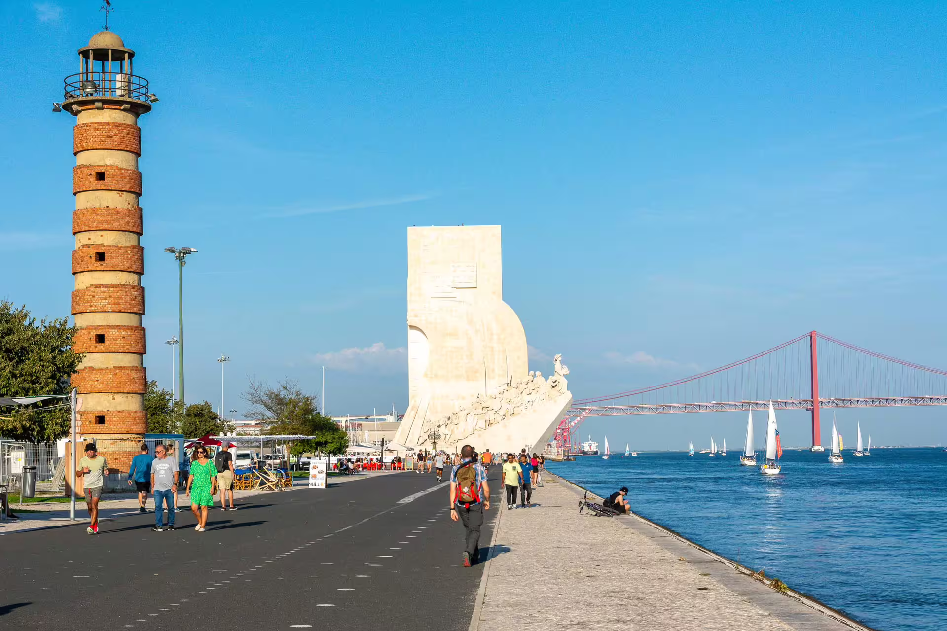 People enjoy a scenic walk along the Tagus River in Belém, Lisbon, showcasing the Monument of Discoveries and 25th April Bridge.